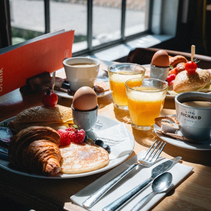 Ontbijtbord met een vegetarisch belegd broodje, croissant, pannenkoekje met vers fruit, gekookt ei, glas verse jus d'orange en een kop koffie op een sfeervol gedekte tafel
